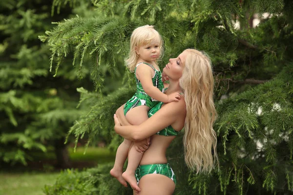 Summer vacations family look, blond girls parenthood portrait. Beautiful Mother holding her little daughter, wears in fashion green swimwear having fun at park.