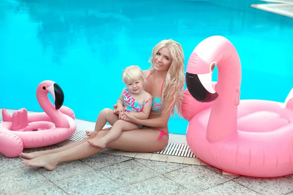 Summer family vacation. Fashion look blond girls portrait. Beautiful Mother holding her little daughter, wears in swim wear posing with Inflatable Flamingo Pool Float by swimming pool on resort.