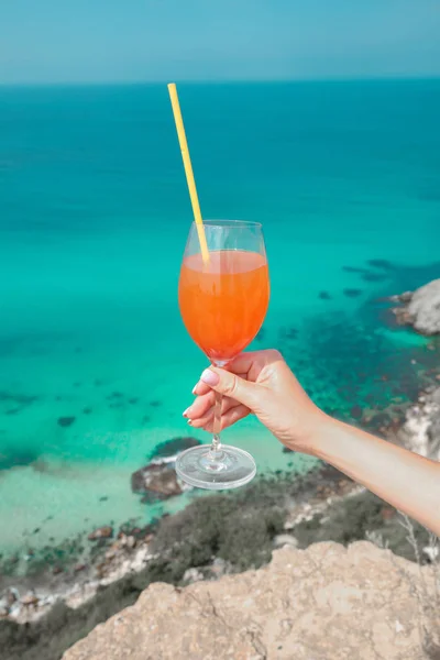 Cheers! Cropped image of Hand with orange coctail glass over turquoise lagoon exotic bay. Lifestyle summer vacation concept. Black sea coast, Crimea.