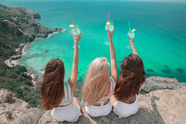 Cheers! Back view of Beautiful traveller girl friends having fun with coctail glass on cliff mountains enjoying lagoon bay over Fiolent, Crimea. Three young women resting on a beach. Travel lifestyle.