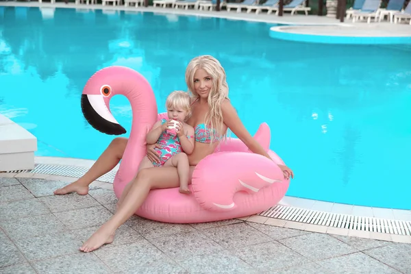Beauty fashion mother with daughter family look. Beautiful blond woman with having fun with little pretty gil wears in swim wear posing on Inflatable Flamingo Pool Float by swimming pool.