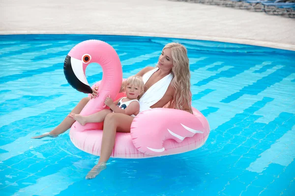 Beauty fashion mother with daughter family look. Beautiful blond woman with having fun with little pretty gil wears in swim wear posing on Inflatable Flamingo Pool Float in swimming pool.