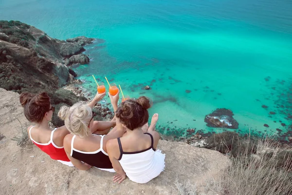 Cheers! Back view of Beautiful traveller girl friends having fun with coctail glass on cliff mountains enjoying lagoon bay over Fiolent, Crimea. Three young women resting on a beach. Travel lifestyle.