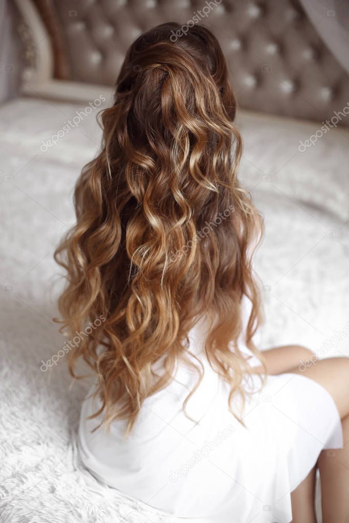 Back view of wedding hairstyle. Beautiful brunette Bride Portrait with curly hair style wearing in white sexy boudoir dressing gown posing on bed in white bedroom.