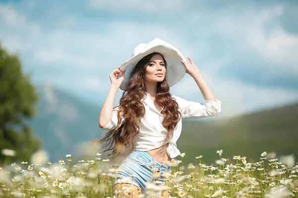 カモミール フィールドの上の白い帽子の美しい少女 屋外自然の中で楽しく健康的なウェーブのかかった髪と屈託のない幸せなブルネット女 の自由スタイル — ストック写真