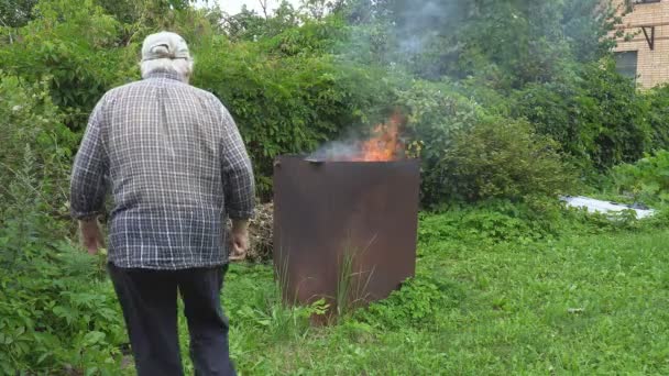 El anciano carga la estufa de jardín, adaptada de un gran tanque de metal, con ramas de arbustos y árboles después de la poda, para que el jardín esté limpio — Vídeos de Stock