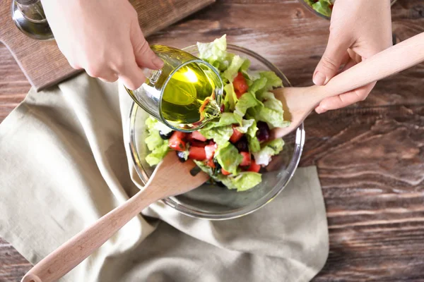 Woman pouring olive oil into bowl with fresh vegetable salad on table