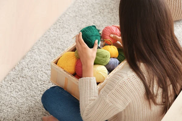 Young woman sitting on carpet and choosing color of knitting threads indoors