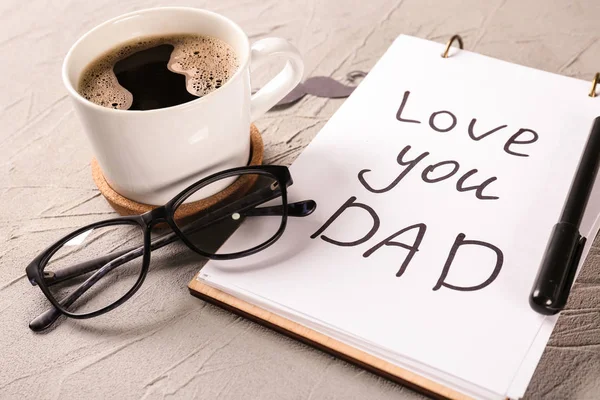 Notebook with LOVE YOU DAD inscription, cup of coffee and glasses on grey textured background. Happy Father\'s Day celebration