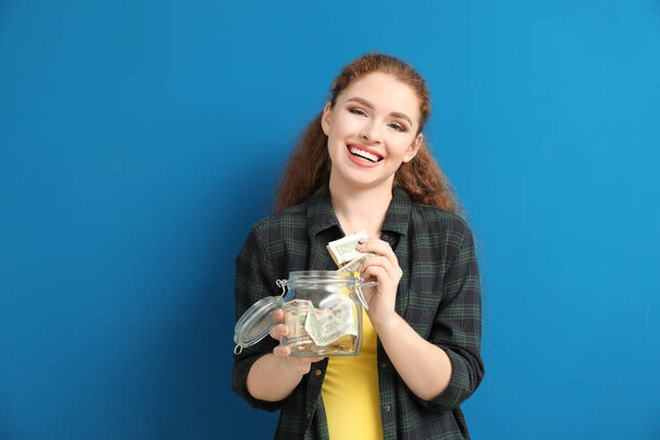 Молодая женщина кладет банкноту в стеклянную банку с деньгами на цветном фоне. Концепция экономии
