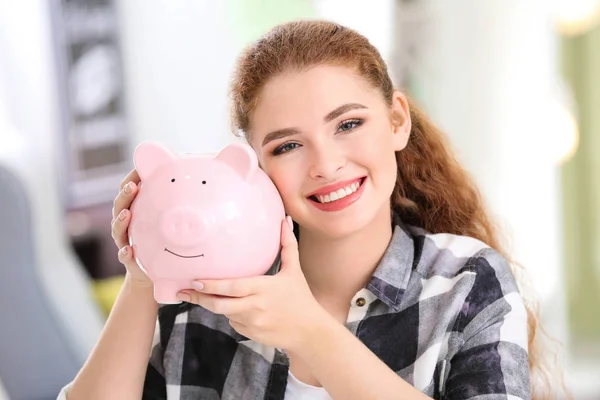 Young woman holding piggy bank indoors. Money savings concept