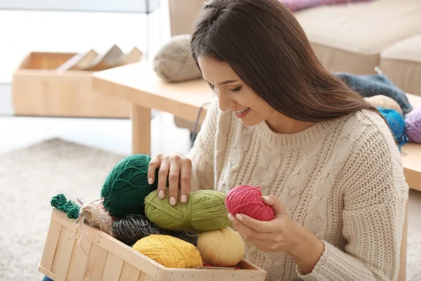 Young woman sitting on carpet and choosing color of knitting threads indoors