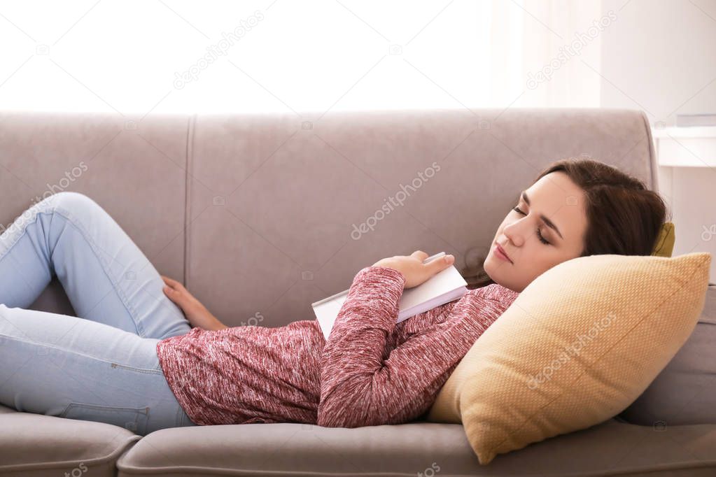 Tired young woman with book sleeping on sofa at home