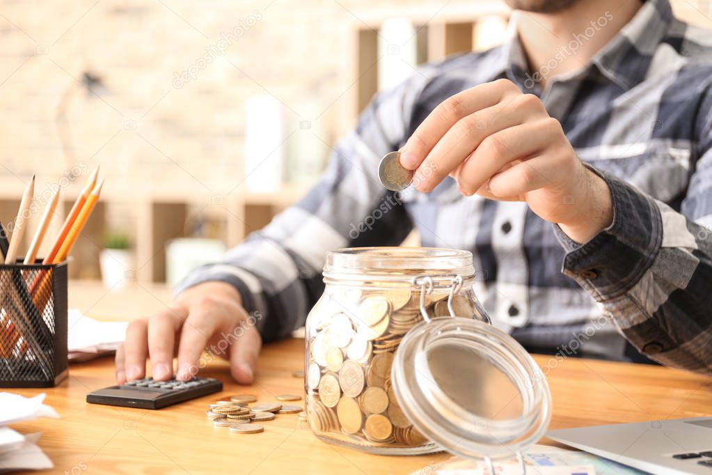 Man putting coin into glass jar indoors. Money savings concept