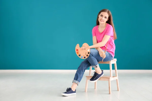 Beautiful female artist sitting on stool against color wall