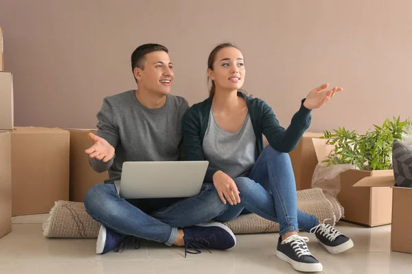 Young couple with laptop sitting on floor near boxes indoors. Moving into new house