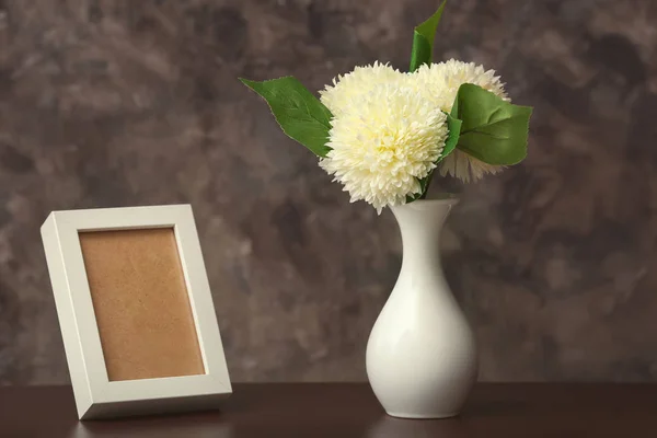 Vase with beautiful blooming flowers and empty frame on table against grey background