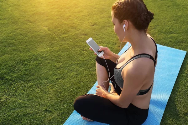 Beautiful young woman sitting on yoga mat and listening to music outdoors