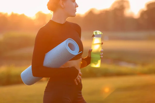 Beautiful young woman with bottle of water and yoga mat outdoors in the morning