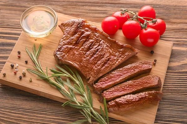 Board with tasty cut steak, rosemary, oil,  tomatoes and spices on wooden background