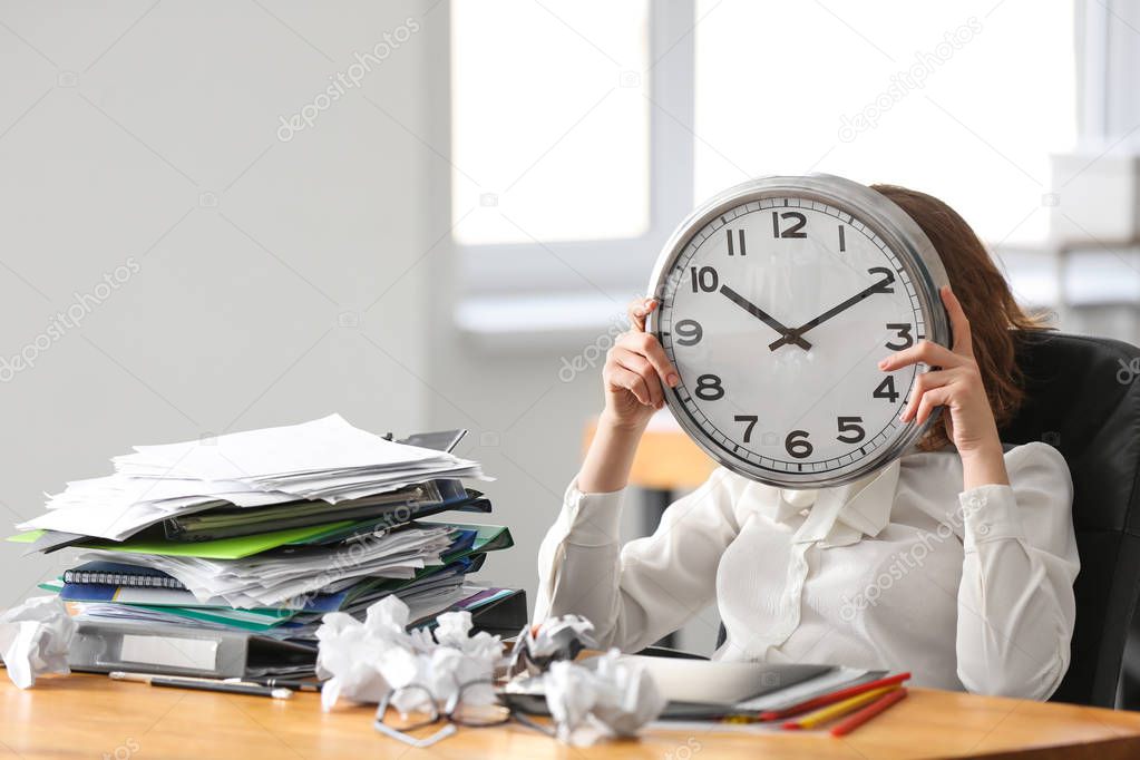 Tired woman hiding face behind clock at table in office. Time management concept