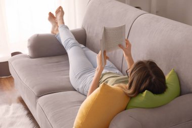 Evdeki kanepede kitap okuyan genç bir kadın.