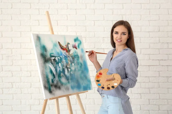 Young female artist with drawing easel against white brick wall