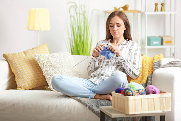 Young woman sitting on sofa and knitting warm sweater at home