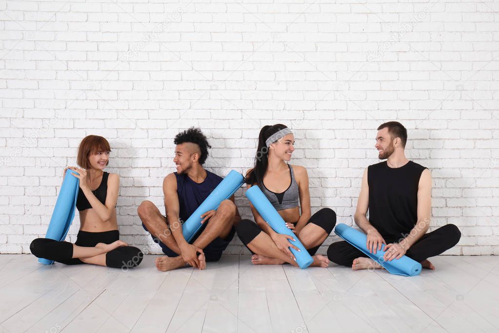 Group of sporty people with yoga mats sitting near white brick wall