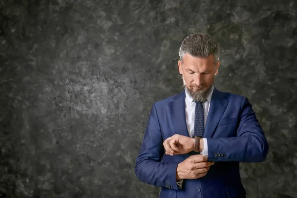 Handsome mature man in elegant suit wearing wristwatch on dark textured background