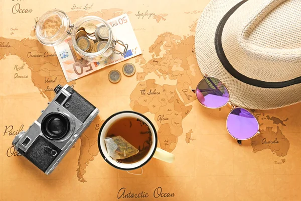 Composition with photo camera, hat, sunglasses, cup of tea and money on vintage world map. Travel planning concept