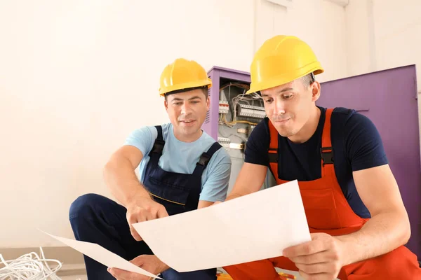 Two electricians with house plan near distribution board indoors