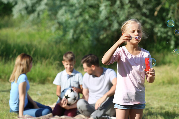 Маленькая девочка раздувает мыльные пузыри, отдыхая со своей семьей в парке

