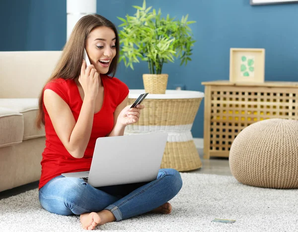 Young woman with laptop and credit card talking on mobile phone at home. Online shopping