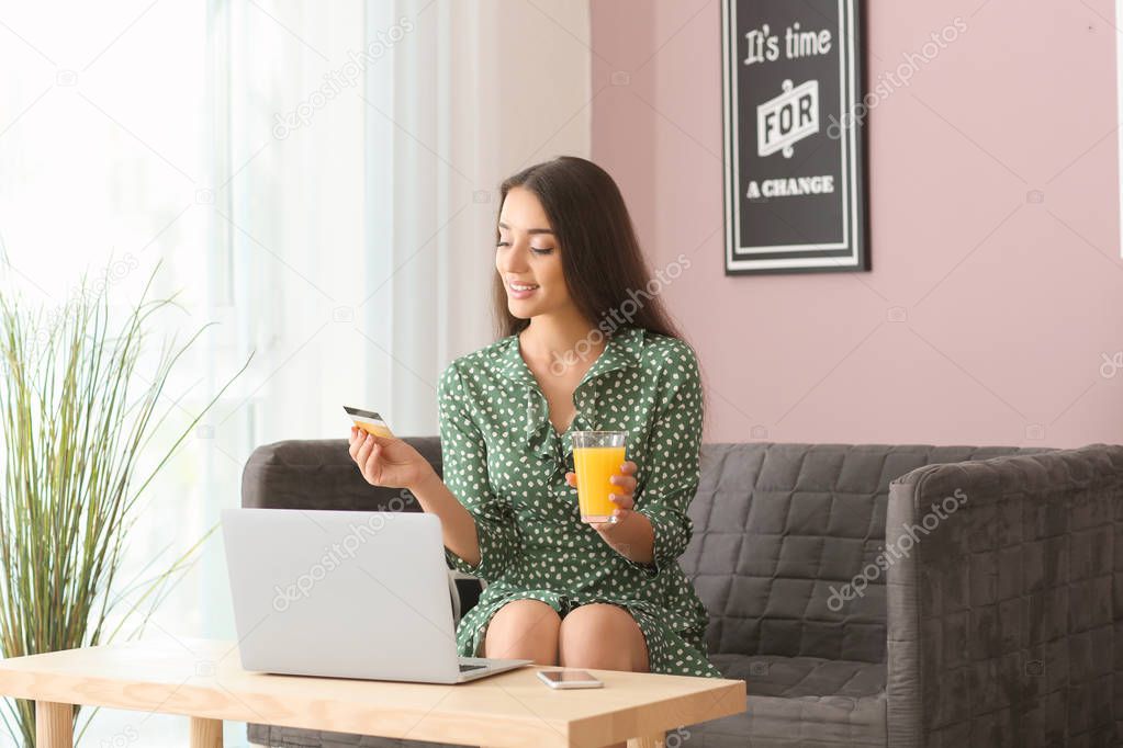 Young woman with credit card and glass of juice sitting on sofa at home. Online shopping
