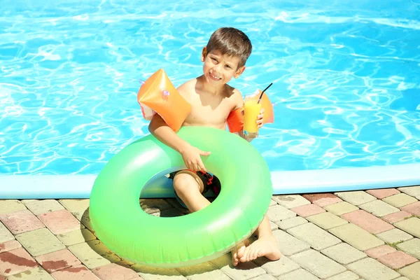 Cute little boy with inflatable ring and glass of juice resting near swimming pool