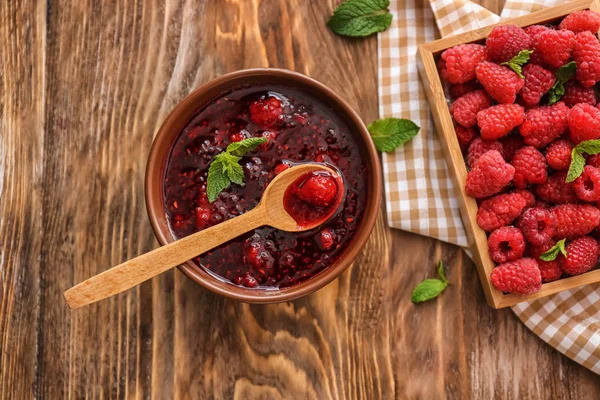 Bowl of sweet jam and box with fresh raspberries on wooden table