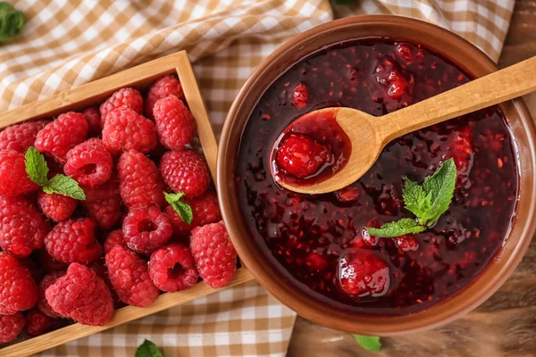 Bowl of sweet jam and box with fresh raspberries on wooden table