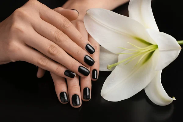 Woman with stylish black manicure and  lily flower on dark background, closeup