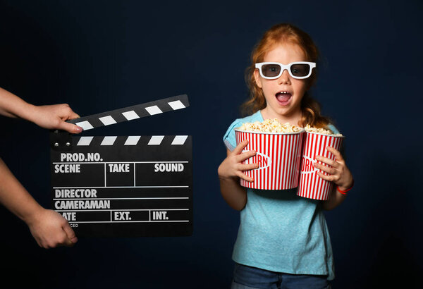 Female hands with clapper board and cute girl holding cups of popcorn on dark background