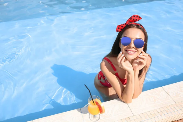 Beautiful young woman with cocktail in swimming pool on summer day
