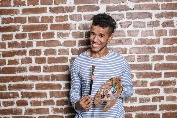 Young African-American artist with palette and brush against brick wall