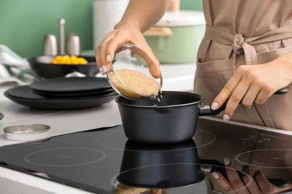 Woman pouring rice into saucepan with boiling water on stove in kitchen