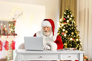 Noel için dekore edilmiş odada dizüstü bilgisayarlı Noel Baba oturuyor.