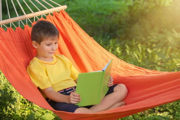 stock image Cute little boy reading book while relaxing in hammock outdoors