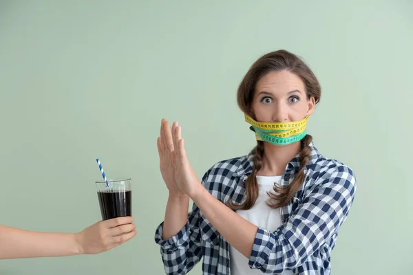 Woman with measuring tapes around her mouth refusing to drink unhealthy beverage on color background. Diet concept