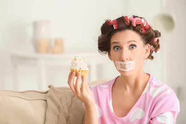 Emotional woman with taped mouth holding tasty tartlet at home. Diet concept