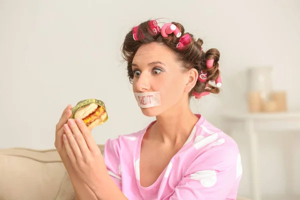 Emotional woman with taped mouth and tasty burger at home. Diet concept
