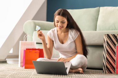 Genç bir kadın evde kredi kartı ve laptop ile online alışveriş yapıyor.