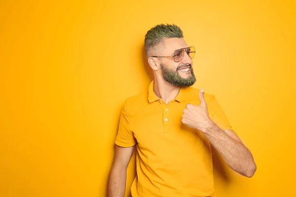 Portrait of handsome man with dyed hair and beard showing thumb-up gesture on color background